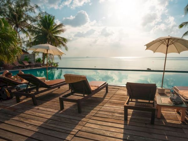 Wooden deck with lounge chairs and umbrellas overlooks a serene infinity pool and ocean, surrounded by palm trees under a bright sky.