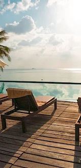 Poolside deck overlooking the ocean, with lounge chairs, umbrellas, and palm trees. The sun is shining through scattered clouds.