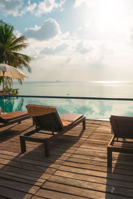 A tranquil poolside scene with lounge chairs, umbrellas, and palm trees overlooking a calm ocean under a partly cloudy sky.