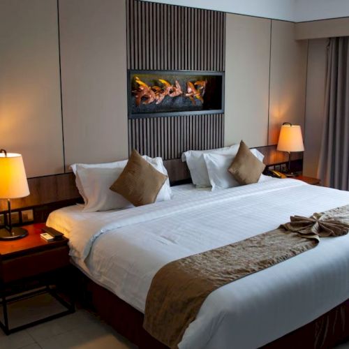 Luxurious hotel room with a large bed, decorative pillows, soft lighting from lamps, and a modern fireplace on the wall.