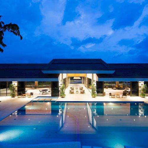 A luxurious building with a large illuminated pool and seating areas, set against a twilight sky, creates a serene and inviting atmosphere.