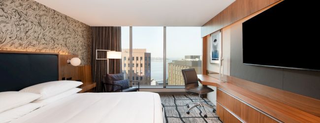 A modern hotel room with a large bed, wall art, desk, chair, TV, and window with a city view.