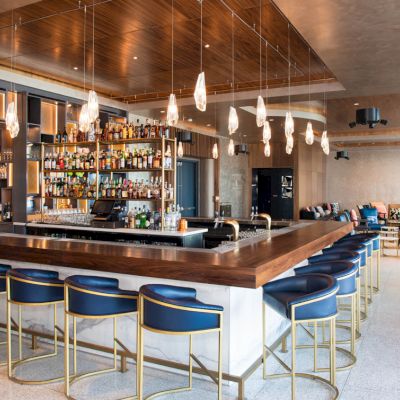 A modern bar with blue stools, hanging lights, and a fully stocked bar counter. The room features a stylish and open seating area.