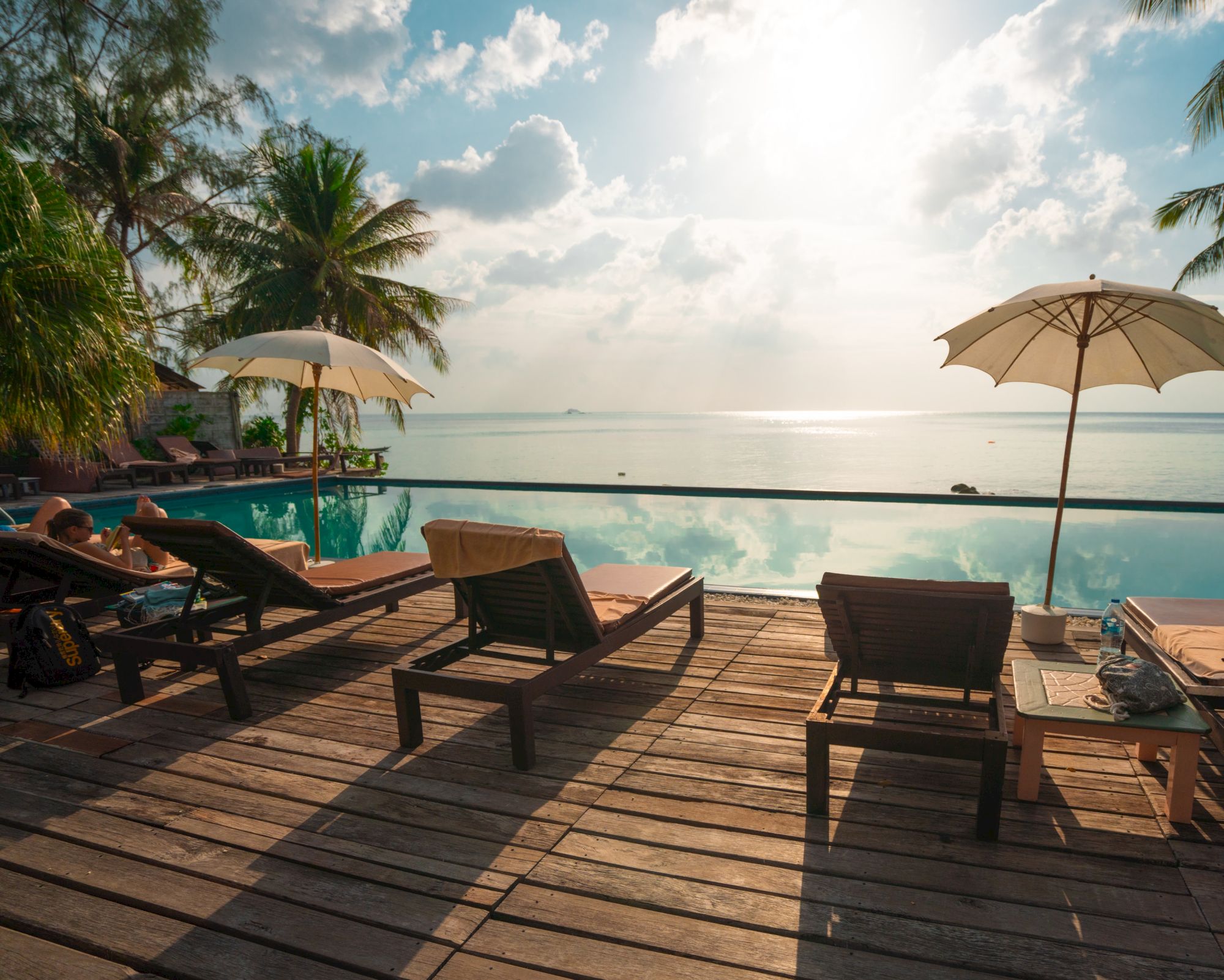 A serene poolside view with lounge chairs, umbrellas, and palm trees, overlooking a tranquil sea under the sunlit sky.