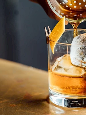 A cocktail is being poured into a glass with ice and a lemon twist garnish, placed on a wooden surface.