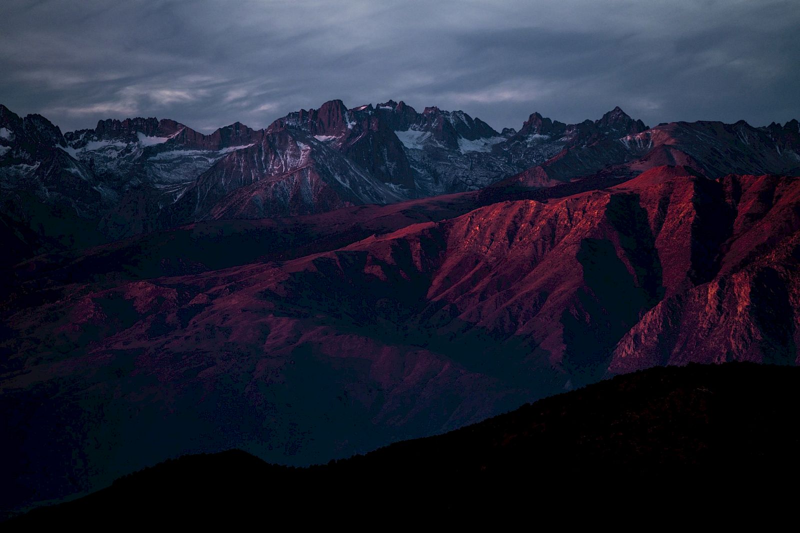 A dramatic mountain range at dusk, with rocky peaks and slopes illuminated by a deep reddish hue under a cloudy sky.