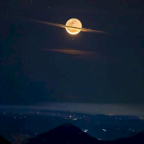 A bright full moon appears with a ring of clouds, resembling Saturn, over a dark landscape with scattered lights below.