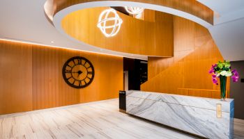 The image shows a modern lobby with a marble desk, spiral wooden ceiling, decorative clock, and floral arrangement.