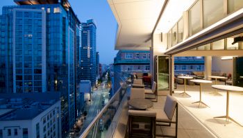A modern urban balcony at dusk overlooks a city street, featuring outdoor seating and a sleek office space inside.