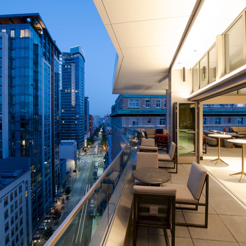 The image shows a modern cityscape at dusk with high-rise buildings and a stylish outdoor terrace featuring chairs and tables.