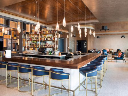 A modern bar with blue stools, pendant lights, stocked shelves, and seating area in a sleek, spacious interior.