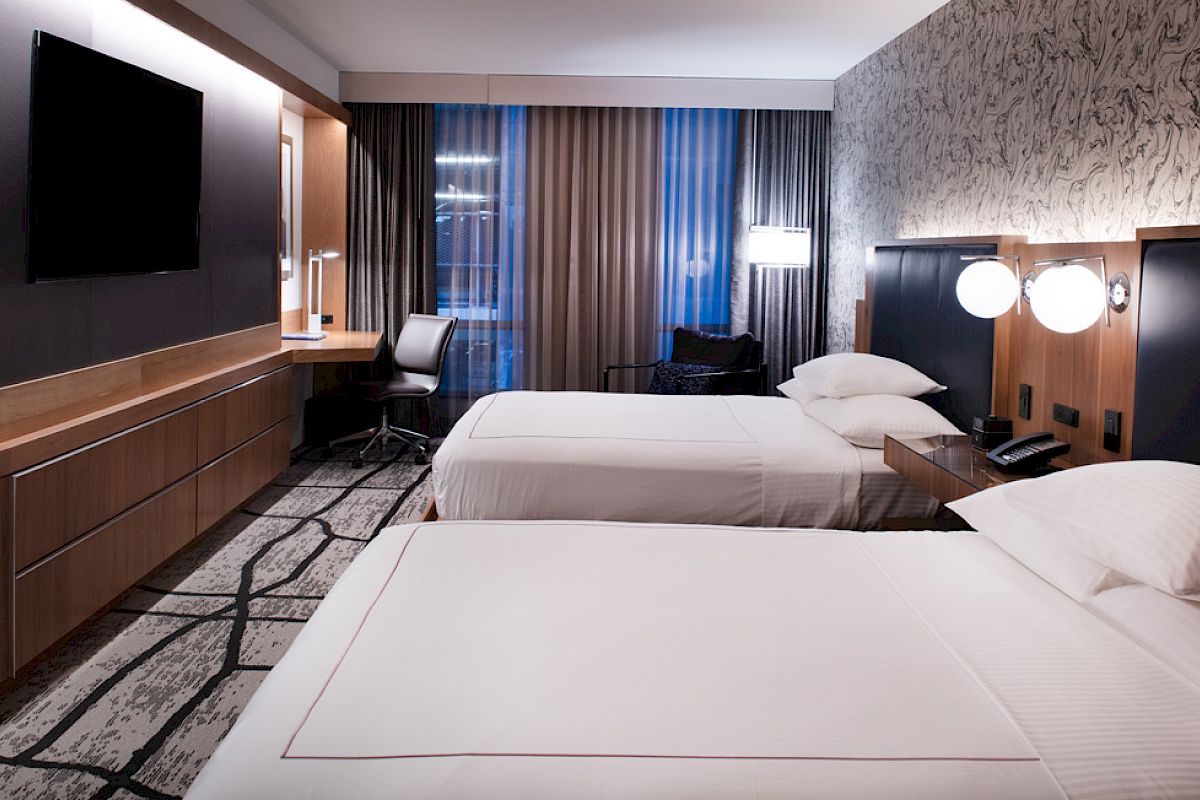 A modern hotel room with two beds, a TV, a desk, and ambient lighting. The decor is sleek and contemporary.
