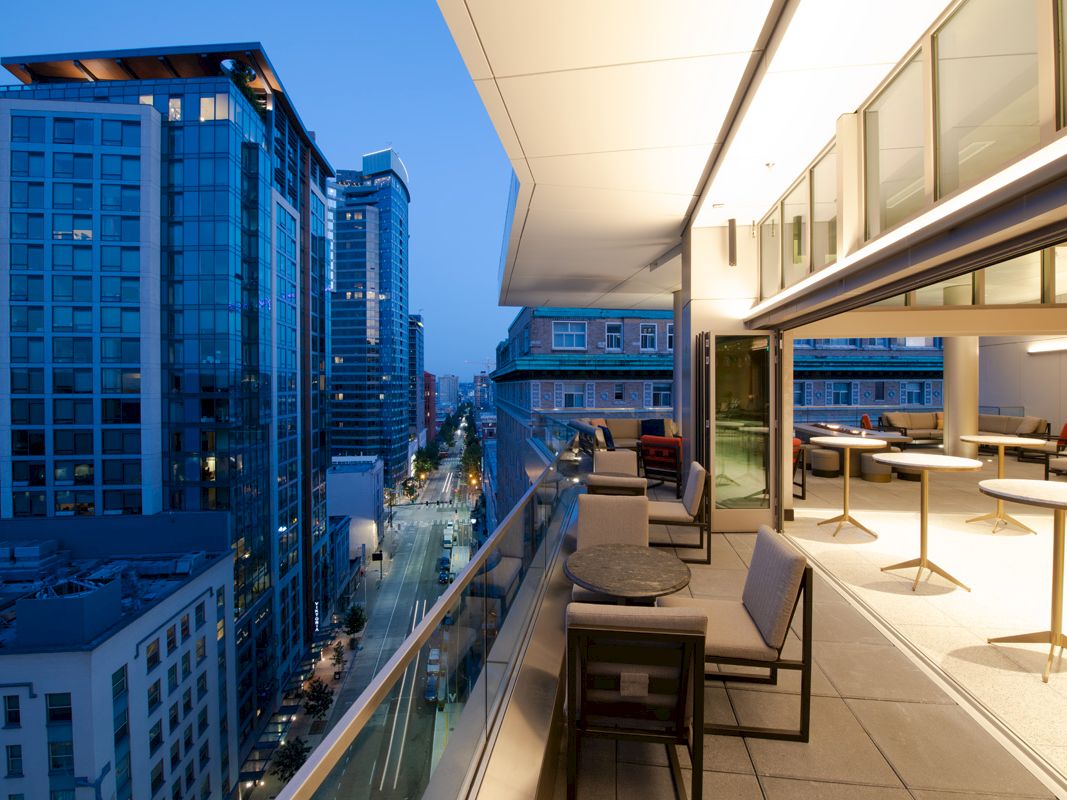 A modern cityscape at dusk features a balcony with seating, overlooking tall buildings and a street below.