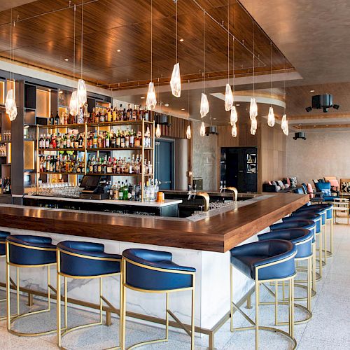 A modern bar with blue stools, hanging lights, and a fully stocked back shelf, set in a spacious lounge area.