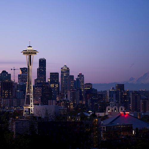 Seattle skyline at dusk featuring the Space Needle and Mount Rainier in the background, with city lights beginning to illuminate.