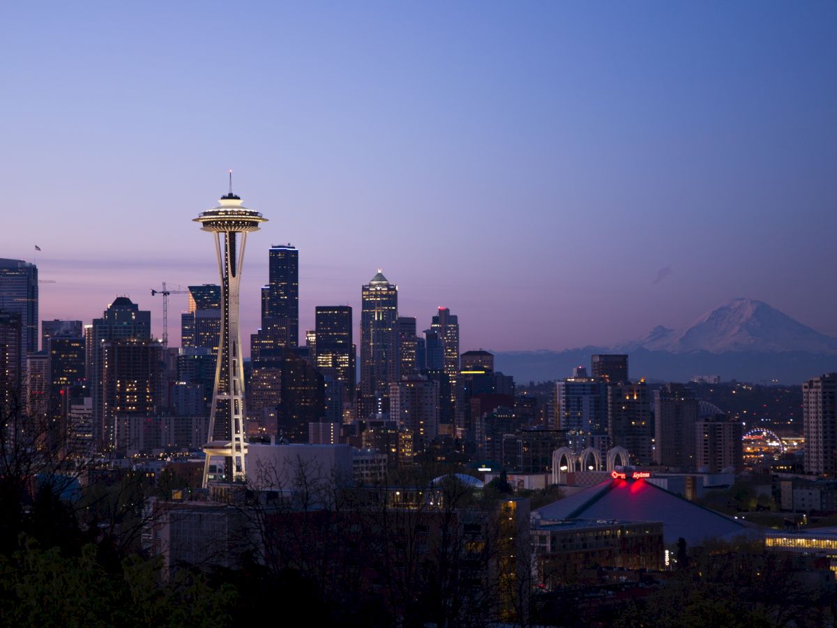 A city skyline at dusk featuring the Space Needle with Mount Rainier visible in the background under a clear sky.