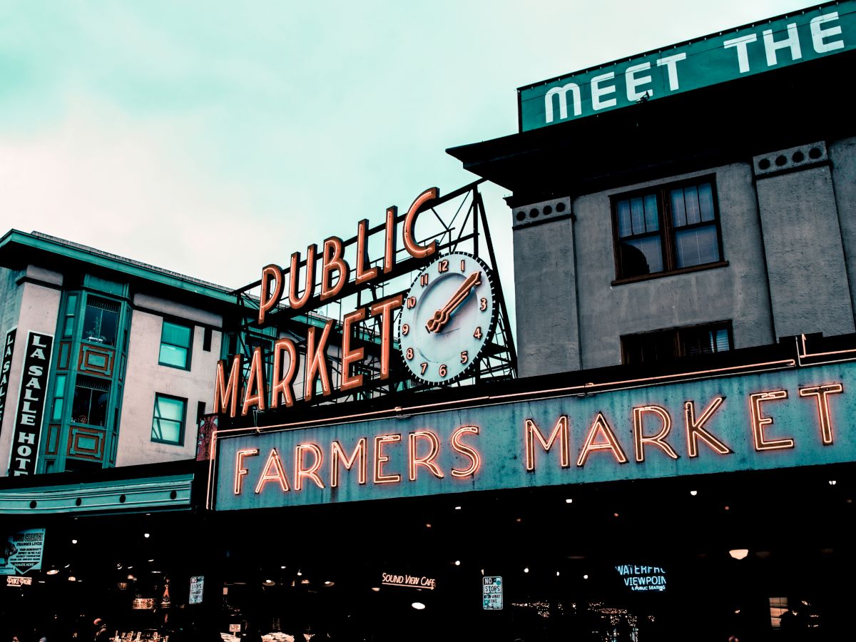 The image shows a famous farmers market entrance with neon signs and a clock, set against a backdrop of buildings and an overcast sky.