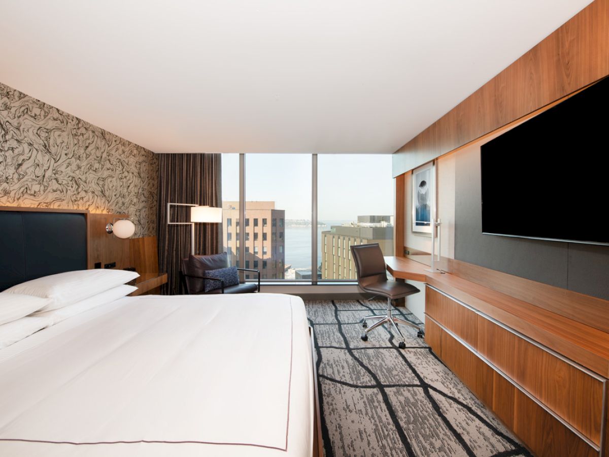 A modern hotel room features a bed, wall-mounted TV, desk with a chair, and a large window with a city view.