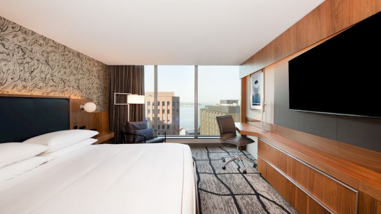 A modern hotel room features a bed, wall-mounted TV, desk with a chair, and a large window with a city view.