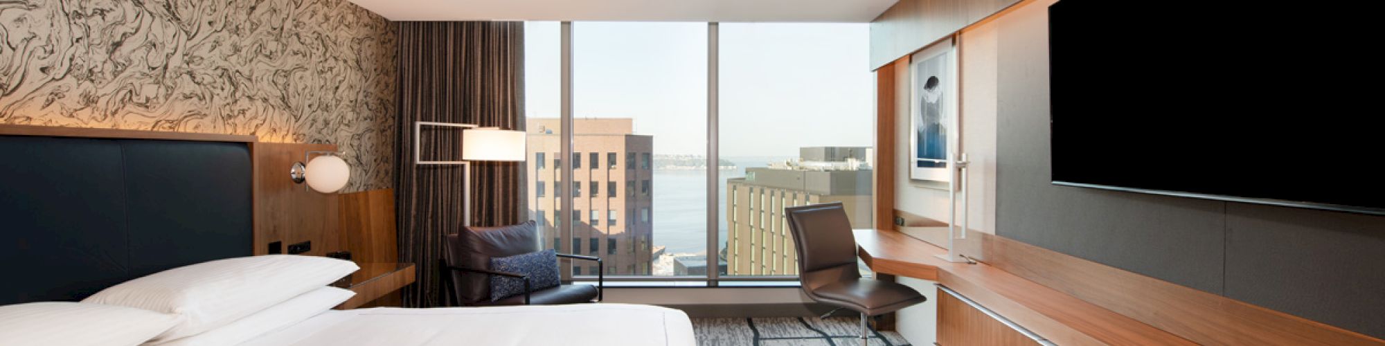 A modern hotel room with a large bed, desk, chair, TV, and city view through large windows.